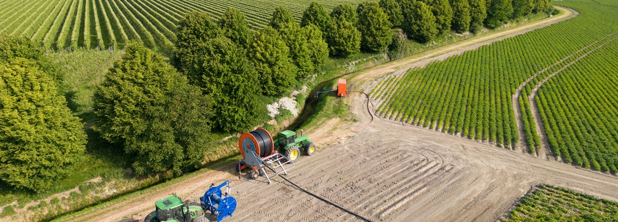 Luchtfoto van een landbouwgebied waar een beregeningsinstallatie wordt opgezet. Twee tractors zijn bezig met het uitrollen van een grote haspel met slang voor irrigatie. De akkers zijn omgeven door bomen en in de verte is een boerderij te zien.