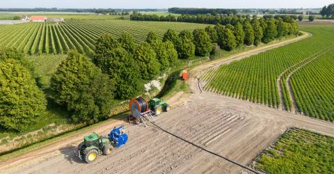 Luchtfoto van een landbouwgebied waar een beregeningsinstallatie wordt opgezet. Twee tractors zijn bezig met het uitrollen van een grote haspel met slang voor irrigatie. De akkers zijn omgeven door bomen en in de verte is een boerderij te zien.